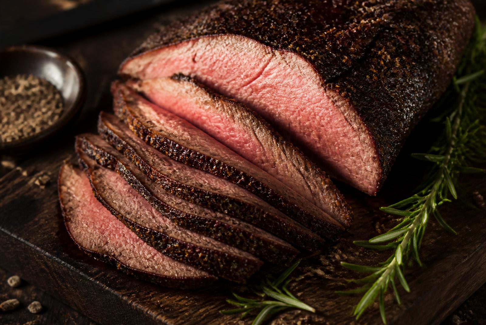 Sliced coffee-crusted tri tip on a dark cutting board with rosemary and cracked pepper